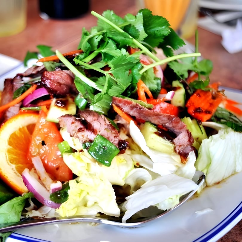 Close-up of a Thai beef salad with lettuce, red onion, orange, and cilantro on a white plate.